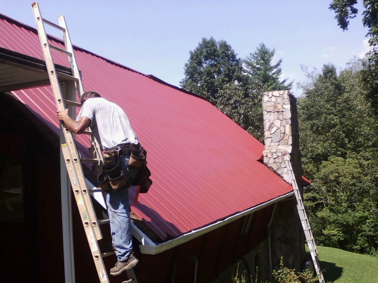 Roofer on a ladder beside a red metal roof and stone chimney — Soffit & Fascia Repair by Roofing Brothers in Saint Paul